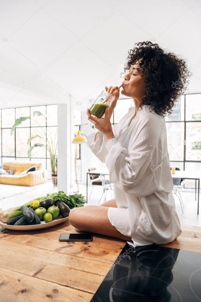 Young african american woman drinking green juice with reusable bamboo straw on kitchen counter. Vertical. Home concept. Healthy lifestyle concept. Copy space