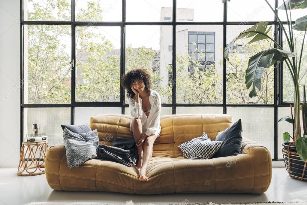 Young and beautiful african american woman with curly hair sitting on a yellow couch looking at camera in a bright loft apartment. Copy space.