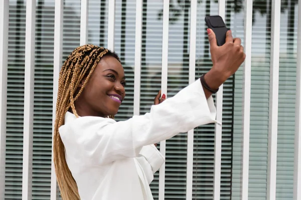 Afroamerikansk ung kvinna ler med en selfie med mobiltelefon. Blonda flätor och vit blazer. — Stockfoto