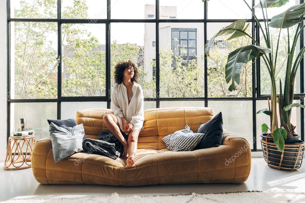 Young and pretty african american woman relaxing and sunbathing on sofa in a bright loft living room.