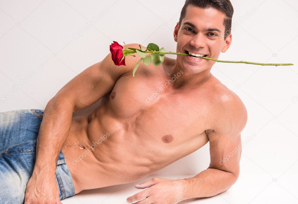 Young muscular man with perfect torso is lying and holding single rose, on white background.