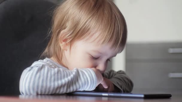 Een klein kind van 2-3 jaar zit op een stoel aan de tafel, kijkt naar de tablet en tikt op het tabletscherm — Stockvideo