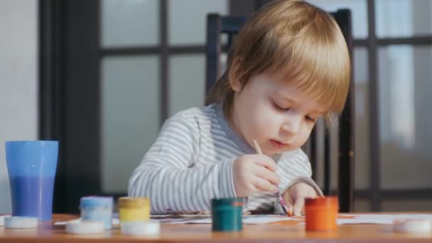 Ένα μικρό αστείο παιδί ηλικίας 2-3 ετών που ζωγραφίζει με μπογιές και βούρτσες, κάθεται σε ένα τραπέζι. Πρόωρη ανάπτυξη παιδιών — Αρχείο Βίντεο
