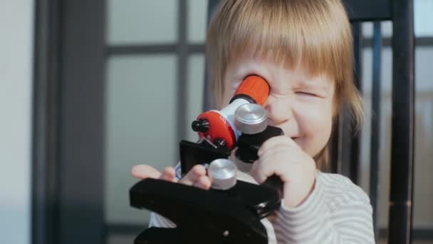 Funny toddler holding toy microscope in hands, squinting and looking into microscope with one eye. — Stock Video