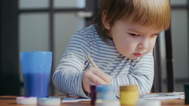 Ein süßer kleiner Junge zeichnet mit Pinsel und Gouache mit Farben auf ein Blatt Papier, das an einem Tisch sitzt. — Stockvideo