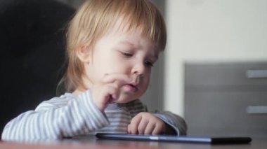 Bir çocuğun portresi masada oturuyor ve hevesle dijital tabletin ekranına bakıyor ve dokunuyor. Çocukların gelişimi için modern elektronik cihazlar.