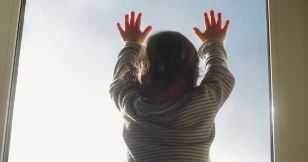 Zadní pohled na dítě stojící u okna a dívající se z okna na modrou oblohu. Dítě se dívá z okna s rukama na skle. Sebeizolace během karantény, covid-19 — Stock video