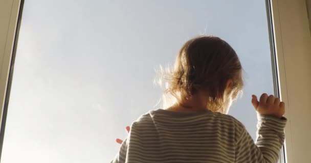 Kleines Kind steht vor dem Fenster und blickt in den blauen Himmel in den Strahlen der untergehenden Sonne und klopft mit den Händen auf Fensterscheiben auf der Suche nach einem Ausgang — Stockvideo