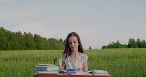 Éducation en plein air. La fille d'âge scolaire s'assied au bureau et regarde des livres dans le pays ouvert. — Video