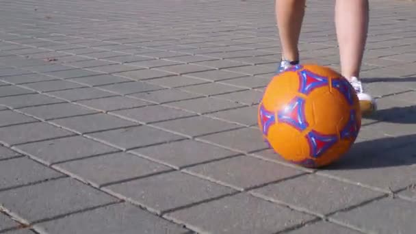 Close-up meninos pés chutando bola de futebol laranja na superfície de pedra asfalto na cidade. — Vídeo de Stock