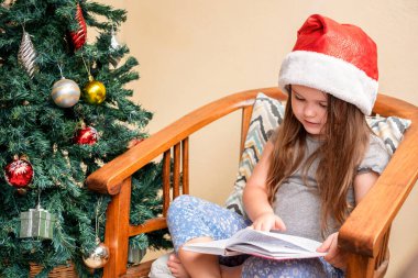 Kız Noel ağacının yanında Noel Baba şapkasında oturuyor. Kitap okuyan bir çocuk. Noel zamanı