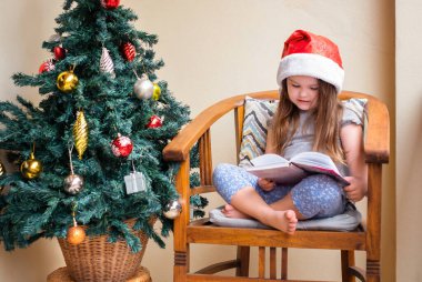 Noel Baba şapkalı mutlu küçük kız Noel ağacının yanındaki sandalyede otururken kitap okuyor.