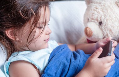 Elinde oyuncak olan bir çocuk sandalyede uzanıyor ve telefonda oynuyor.