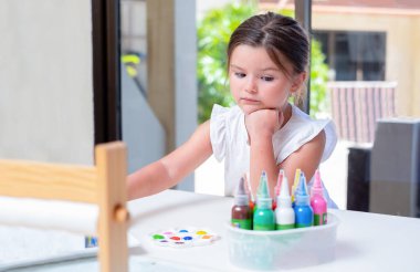 Çocuk boyalara bakmaktadır. Yaratıcılık takımıyla masada oturan kız.