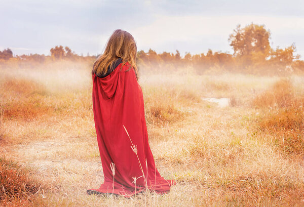A child in a red cloak stands in a field in autumn