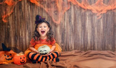 Cadı gibi giyinmiş çocuk gülüyor ve elinde Cadılar Bayramı süslemeleri olan bir balkabağı tutuyor.