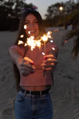 Bir kadın maytap tutuyor ve gülümsüyor. İmajın mutlu ve neşeli bir havası var. Kadın kıyafeti rahat ve rahat, kot pantolon ve pembe gömlekli.