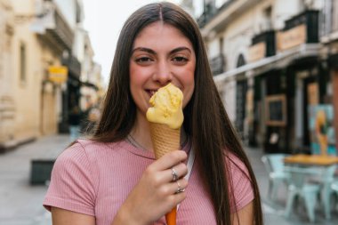 Bir kadın elinde dondurma külahı tutuyor. Gülümsüyor ve ziyafetinin tadını çıkarıyor. Sahne, birkaç sandalye ve masa olan bir sokakta geçiyor.