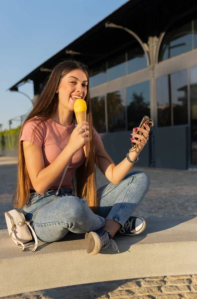 Neşeli bir kadın sokakta oturmuş dondurma külahı yiyor, kameraya bakıyor, diğer elinde bir cep telefonu tutuyor..