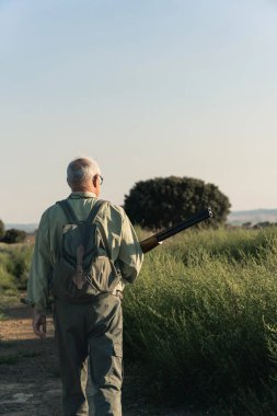 Yeşil bir tarlada toprak bir yolda yürüyen, bir av tüfeği ve sırt çantası taşıyan yaşlı bir adam.