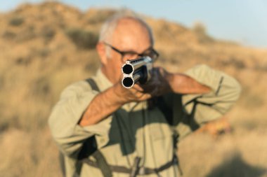Üst düzey avcı elinde bir av tüfeği tutuyor, düz nişan alıyor, arka planda.