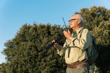 Dışarıda avlanan yaşlı bir adam, elinde pompalı tüfek ve telsizle iletişim kuruyor.