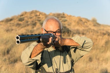 Yaşlı bir adam av tüfeğiyle, kurak kırsal bölgede avlanırken eğleniyor.
