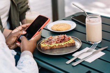 Kadınlar açık bir kafede akıllı telefon kullanırken avokado tostu ve içeceklerle brunch yapıyorlar.