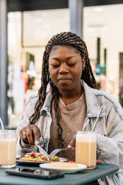 Örgülü saçlı bir kadın avokado tostu yiyor ve açık hava masasında içiyor.