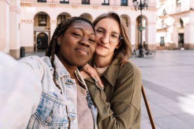 Genç ve çeşitli kadınlar gülümsüyor ve kucaklaşıyorlar. Şehir meydanında selfie çekiyorlar.