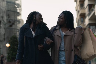İki neşeli Afrikalı kadın kol kola gezip birbirlerine bakıyorlar şehrin sokaklarında suç ortaklığıyla, birlikte alışveriş gününün tadını çıkarıyorlar..