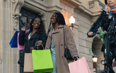 İki arkadaş alışveriş torbalarını şehir caddesinde taşımaktan mutlu olurlar.
