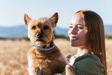 Gülümseyen kızıl saçlı kadın altın buğday tarlasında sevimli köpeğiyle eğleniyor. Yürek ısıtan bir arkadaşlık ve doğa manzarası yaratıyor.