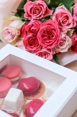 Bir buket pembe gülün arka planında çeşitli kremalı kekler ve beyaz bir kutuda kurabiyeler.