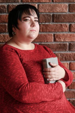 Kırmızı elbiseli kısa saçlı esmer, elinde kırmızı tuğlalı bir arka planda kitap tutuyor.