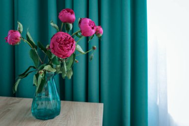 Masanın üzerinde mavi ve beyaz perdelerin arkasında duran mavi cam vazolu bir buket kırmızı şakayık.