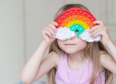 Sarışın bir kız elinde gökkuşağı şeklinde bir oyuncak tutuyor ve yüzünü onunla kaplıyor. Mutlu çocuk bir oyuncağın arkasından gözetliyor.