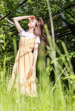 Sarı kareli elbiseli esmer bir kız güneşli bir günde uzun yeşil çimenlerin arasında döner.