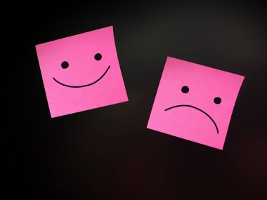 Siyah duvarda üzgün ve mutlu yüzler çizilmiş iki pembe etiket.