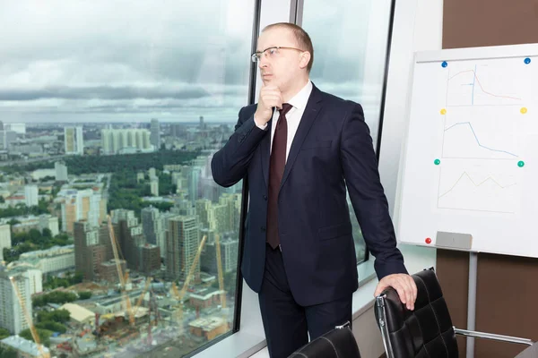 Ofiste pencereye yakın duran, koltuğa uzanıp şehre bakan, takım elbiseli dalgın yöneticinin yan görünüm portresi.