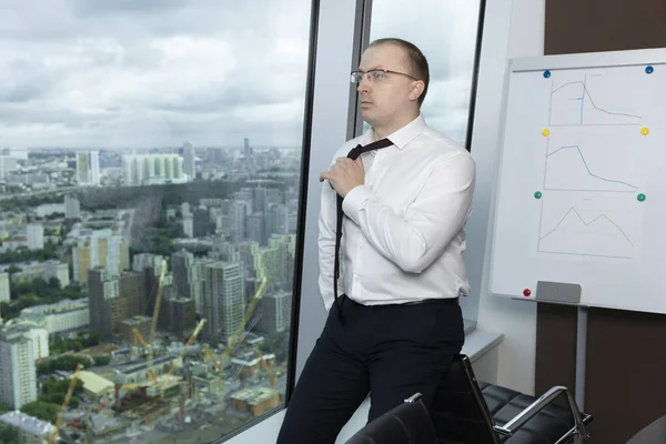 Beyaz gömlekli müdür ofisteki pencerenin yanındaki koltukta oturur ve kravatını çeker ve koparır.