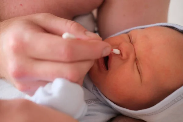 Padre sostiene a su bebé recién nacido en sus brazos y se limpia la nariz con un hisopo de algodón. Retrato de un bebé llorando — Foto de Stock