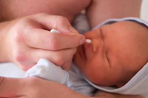 Padre sostiene a su bebé recién nacido en sus brazos y se limpia la nariz con un hisopo de algodón. Retrato de un bebé llorando — Foto de Stock