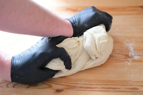 Chef en gants noirs pétrit pâte pour faire du pain, khinkali, boulettes et pâtisseries. Mains masculines sur un fond de table en bois. Concept de cuisson. — Photo