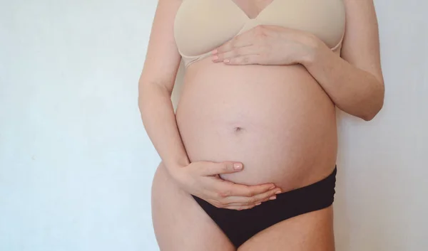 ランジェリー姿の妊婦が白い壁を背景に立ち腹を撫でている コンセプト 母親と妊娠 — ストック写真