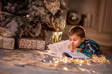 Çocuk süslü bir Noel ağacının yanında yerde yatıyor. Noel hikayeleriyle dolu bir kitap okuyorum. Evdeki sıcak kış akşamları. Noel ve evde yeni yıl tatili kavramı. seçici odak; çevre ışığı; alanın sığ derinliği