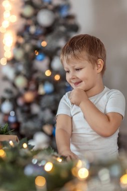 4-5 yaşlarında güzel bir çocuk Noel ağacının yanında oturuyor ya da ayakta duruyor. Tatil ve hediyeler hayal eder, bir dilek tutar. Kış tatili kavramı. Seçici yumuşak odak, bokeh (bulanık arkaplan).