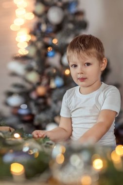 4-5 yaşlarında güzel bir çocuk Noel ağacının yanında oturuyor ya da ayakta duruyor. Tatil ve hediyeler hayal eder, bir dilek tutar. Kış tatili kavramı. Seçici yumuşak odak, bokeh (bulanık arkaplan).