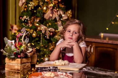 A smiling girl is sitting at a table in the decorated kitchen, with raw dough on the table in front of her. Her nose is smeared with flour. Selective focus. Homemade cakes, Christmas sweets, winter holiday concept.