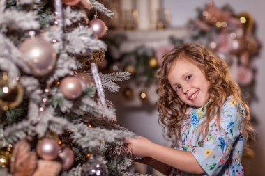 Kıvırcık uzun saçlı güzel bir kız süslü bir Noel ağacının yanında duruyor. Noel ağacını süslüyor. Noel ağacına balon ve oyuncak asar. Evde kış tatili kavramı. Kapat..